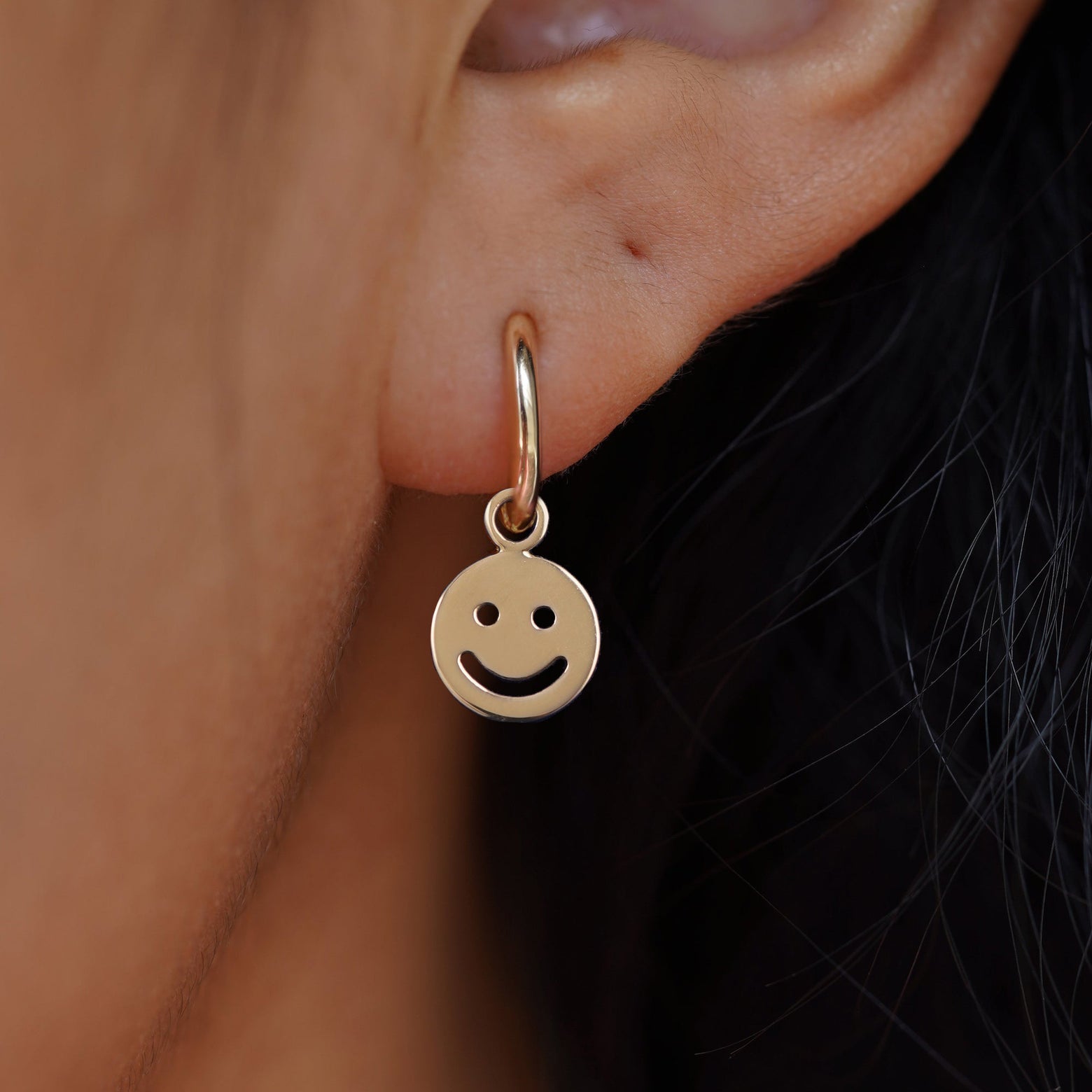 Close up view of a model's ear wearing a yellow gold Smiley Face Charm on a Small Curvy Huggie Hoop
